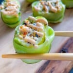 Close-up of chopsticks holding a Shrimp Cucumber Roll wrapped in thin cucumber slices, topped with sauce and sprinkled with sesame seeds and seasoning. More similar sushi rolls are on a wooden board in the background.