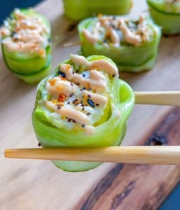 Close-up of chopsticks holding a Shrimp Cucumber Roll wrapped in thin cucumber slices, topped with sauce and sprinkled with sesame seeds and seasoning. More similar sushi rolls are on a wooden board in the background.