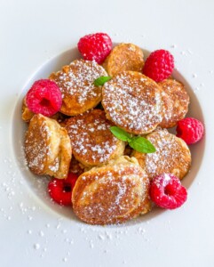 A white plate with small, round pancakes sprinkled with powdered sugar, garnished with fresh raspberries and mint leaves—a charming breakfast treat inspired by the comfort of a Chocolate Banana Bread Recipe.