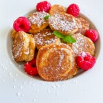 A plate of small golden Gluten Free Banana Pancakes dusted with powdered sugar, garnished with fresh raspberries and mint leaves, on a white plate.