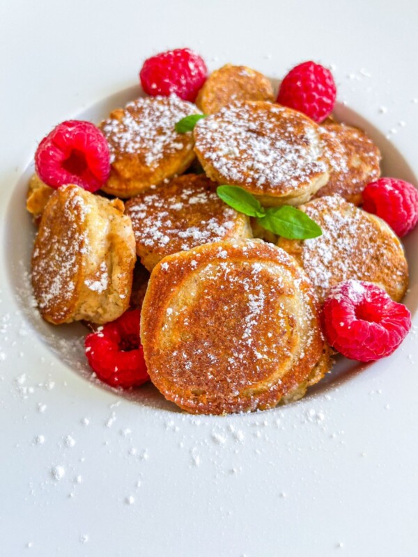 A plate of small golden Gluten Free Banana Pancakes dusted with powdered sugar, garnished with fresh raspberries and mint leaves, on a white plate.