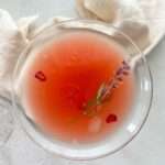 A top-down view of a pink Cosmopolitan Drink with ice, garnished with a sprig of lavender and red pomegranate seeds, set on a light surface with a cream-colored cloth in the background.