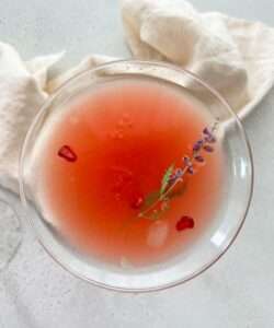 A top-down view of a pink Cosmopolitan Drink with ice, garnished with a sprig of lavender and red pomegranate seeds, set on a light surface with a cream-colored cloth in the background.