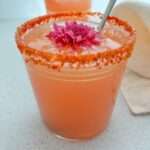 A pink Guava Margarita cocktail in a glass with a chili-salt rim, topped with a pink flower and served with a metal straw, sits on a light surface next to a beige napkin—perfect for trying out your favorite Guava Margarita recipe.