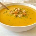 A bowl of creamy orange Cream of Zucchini Soup topped with croutons and sprinkled herbs, with a spoon resting inside.