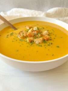 A bowl of creamy orange Cream of Zucchini Soup topped with croutons and sprinkled herbs, with a spoon resting inside.