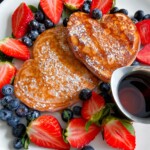 A plate with two heart-shaped Gluten Free Banana Pancakes dusted with powdered sugar, surrounded by fresh strawberries and blueberries, and a small metal container of syrup on the side.