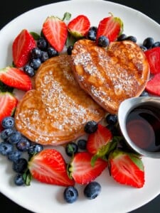 A plate with two heart-shaped Gluten Free Banana Pancakes dusted with powdered sugar, surrounded by fresh strawberries and blueberries, and a small metal container of syrup on the side.
