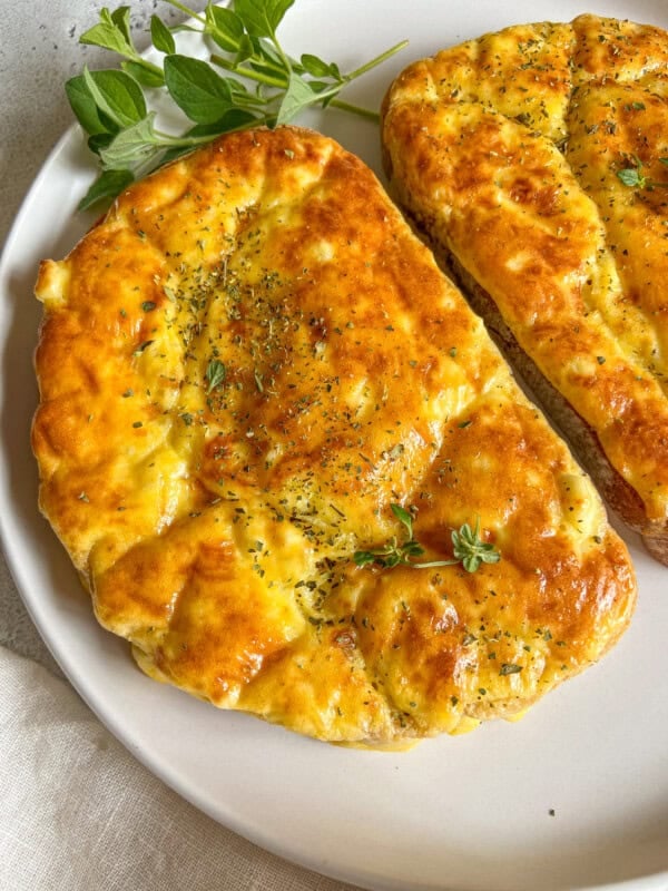 Two pieces of golden, freshly baked flatbread garnished with herbs are served on a white plate, accompanied by a sprig of fresh green oregano, reminiscent of flavors found in Feta Egg Toast.