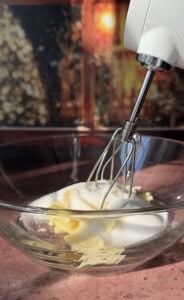 A close-up of a glass bowl containing butter and sugar being mixed for Apple Cookies (Scones) with an electric hand mixer, set against a blurred, warm-toned background.