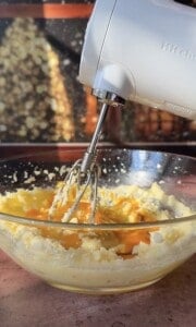 A hand mixer whips together eggs, sugar, and butter in a clear glass bowl, preparing the base for delicious Apple Cookies (Scones), while sunlight streams through a window in the background.