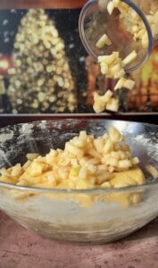 Chopped apples are poured from a cup into dough for delicious Apple Cookies (Scones), with a blurred festive background featuring warm lights and a decorated tree.