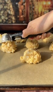 A hand uses a metal scoop to place balls of Apple Cookies (Scones) dough with visible coconut flakes onto a parchment-lined baking sheet. Other dough balls are already arranged on the sheet, ready for baking.