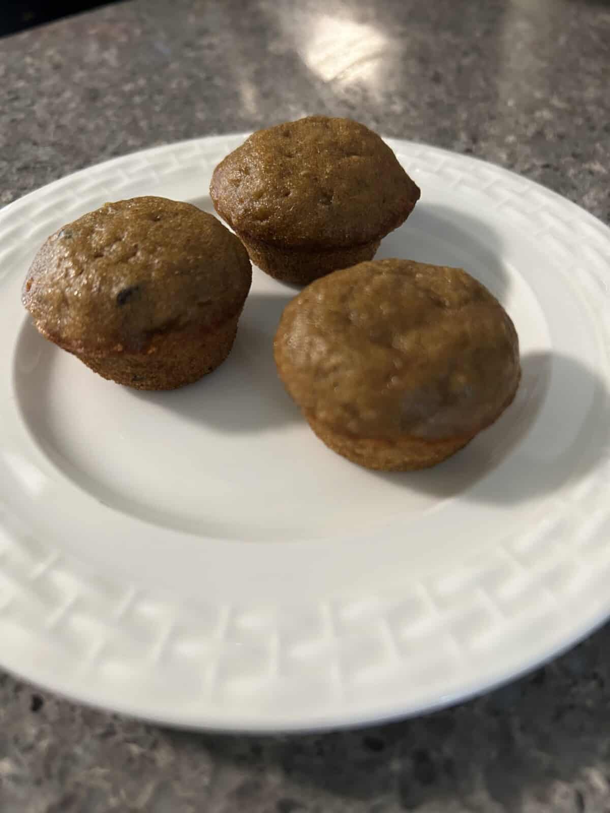 Three small muffins, inspired by the Best Banana Bread Recipe, are arranged on a white plate with a patterned rim, set on a speckled gray countertop.