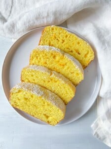 Four slices of Revani Cake, a yellow pound cake topped with powdered sugar, are neatly arranged on a white plate, placed on a white surface with a white cloth in the background.