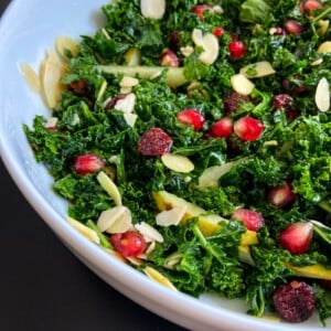 A close-up of a kale salad in a white bowl, inspired by the Jennifer Aniston Salad, featuring pomegranate seeds, dried cranberries, sliced almonds, and thin apple slices.