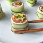 Chopsticks hold a Salmon Cucumber Roll filled with cream cheese and smoked salmon on a white plate, surrounded by similar rolls garnished with chopped green herbs.