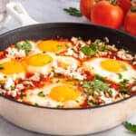 A skillet filled with a classic Shakshouka Recipe, featuring poached eggs in tomato sauce, topped with crumbled cheese and chopped parsley. Fresh tomatoes, parsley, and slices of bread are in the background.