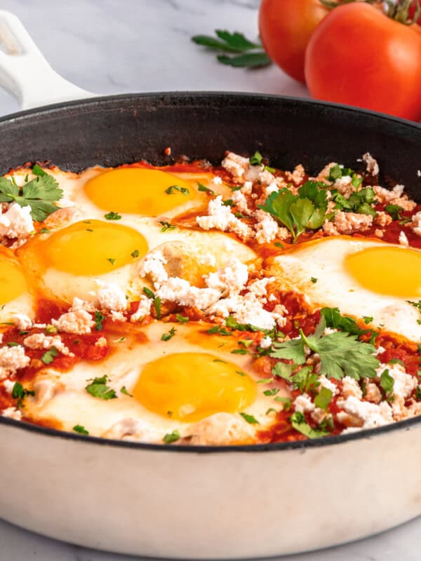 A skillet filled with a classic Shakshouka Recipe, featuring poached eggs in tomato sauce, topped with crumbled cheese and chopped parsley. Fresh tomatoes, parsley, and slices of bread are in the background.