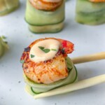 Close-up of seared scallops topped with creamy sauce, served on cucumber ribbons, reminiscent of Shrimp Cucumber Rolls, with one scallop being picked up by chopsticks on a white plate.
