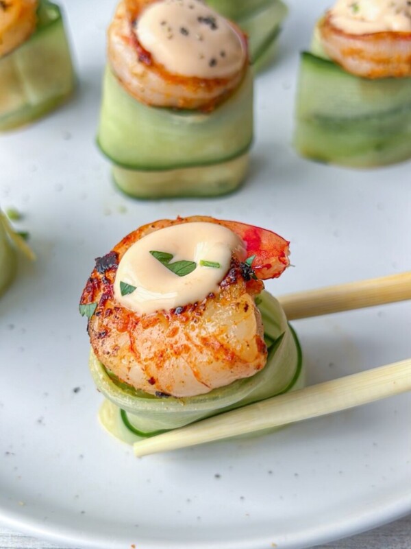 Close-up of seared scallops topped with creamy sauce, served on cucumber ribbons, reminiscent of Shrimp Cucumber Rolls, with one scallop being picked up by chopsticks on a white plate.