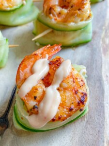 A grilled shrimp topped with creamy sauce rests on a cucumber boat, secured with a toothpick, and served on a wooden surface.