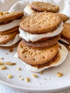A close-up of homemade Oven Baked Smores with toasted marshmallows and chocolate sandwiched between round biscuits, sprinkled with chopped nuts, arranged on a white plate.