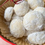 A close-up of several round, white cookies coated in powdered sugar, arranged in a brown paper liner inside a red bowl. Perfect for any Apple Cookies Recipe, with a decorative red string visible in the upper left corner.