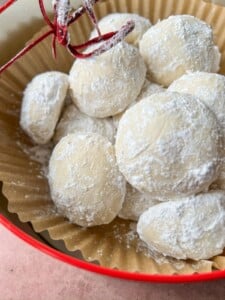 A close-up of several round, white cookies coated in powdered sugar, arranged in a brown paper liner inside a red bowl. Perfect for any Apple Cookies Recipe, with a decorative red string visible in the upper left corner.