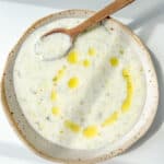 A bowl of creamy Cold Cucumber Soup with herbs, garnished with a drizzle of olive oil in the shape of a smiley face, and a wooden spoon resting in the bowl.
