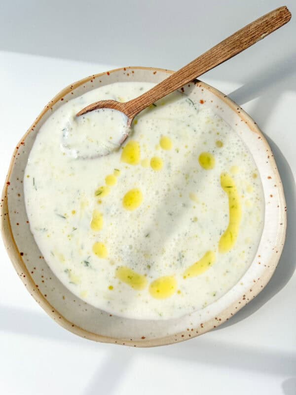 A bowl of creamy Cold Cucumber Soup with herbs, garnished with a drizzle of olive oil in the shape of a smiley face, and a wooden spoon resting in the bowl.