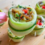 A hand holds a skewer with a zucchini roll-up stuffed with melted cheese, red peppers, and herbs. Other zucchini roll-ups share the wooden surface, offering fresh inspiration for snacks like Cucumber Salmon Boats.