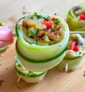 A hand holds a skewer with a zucchini roll-up stuffed with melted cheese, red peppers, and herbs. Other zucchini roll-ups share the wooden surface, offering fresh inspiration for snacks like Cucumber Salmon Boats.