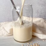 A glass of oat milk is being poured, with a metal straw inside. Some oats are scattered on the surface nearby, and a light cloth is in the background.