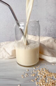 A glass of oat milk is being poured, with a metal straw inside. Some oats are scattered on the surface nearby, and a light cloth is in the background.