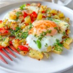 A close-up of two fried eggs served on a bed of sautéed diced potatoes and red bell peppers, garnished with fresh herbs on a white plate with a red fork.
