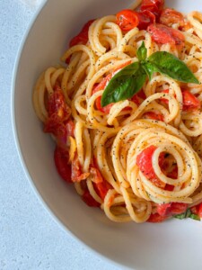 A close-up of a bowl of spaghetti with cherry tomatoes, black pepper, and fresh basil leaves, garnished with a sprinkle of pepper. The pasta is served in a white bowl on a light surface.