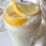 A creamy, frothy lemon drink served in a clear glass, garnished with two fresh lemon slices on top, with a white cloth in the background.
