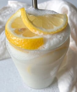 A creamy, frothy lemon drink served in a clear glass, garnished with two fresh lemon slices on top, with a white cloth in the background.