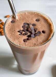 A glass of chocolate smoothie topped with chocolate chips, served with a metal straw. Some whole almonds are scattered in the background on a light surface.