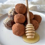 A plate of chocolate truffles dusted with cocoa powder, some coated in chopped nuts, with a honey dipper resting on the plate and honey pooling beneath it.