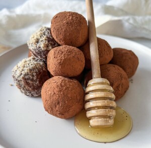 A plate of chocolate truffles dusted with cocoa powder, some coated in chopped nuts, with a honey dipper resting on the plate and honey pooling beneath it.