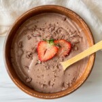 A wooden bowl filled with chocolate smoothie topped with two strawberry slices and chocolate shavings, with a wooden spoon resting inside. A light-colored cloth is in the background.