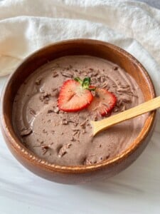A wooden bowl filled with creamy chocolate smoothie, topped with two sliced strawberries and chocolate shavings, with a wooden spoon resting in the bowl on a light surface.