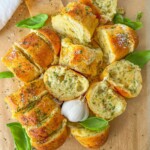 Sliced garlic bread topped with herbs and cheese, arranged on parchment paper with fresh basil leaves and a whole garlic bulb in the center.
