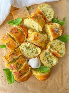 Sliced garlic bread topped with herbs and cheese, arranged on parchment paper with fresh basil leaves and a whole garlic bulb in the center.