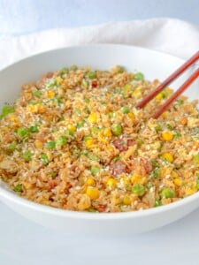 A white bowl filled with delicious vegetable fried rice, including peas, corn, and carrots, garnished with sesame seeds. Try this Vegetable Fried Rice Recipe and enjoy it with chopsticks resting on the side of the bowl.