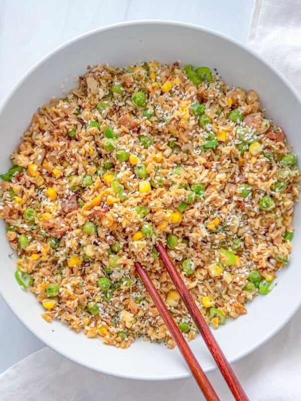 A white bowl filled with vegetable fried rice recipe, mixed with peas, corn, carrots, and garnished with sesame seeds. A pair of wooden chopsticks rests on the edge of the bowl.