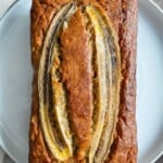 A loaf of banana bread topped with three lengthwise slices of banana sits on a white plate, showcasing its golden-brown crust and moist texture.