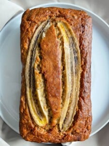 A loaf of banana bread topped with three lengthwise slices of banana sits on a white plate, showcasing its golden-brown crust and moist texture.